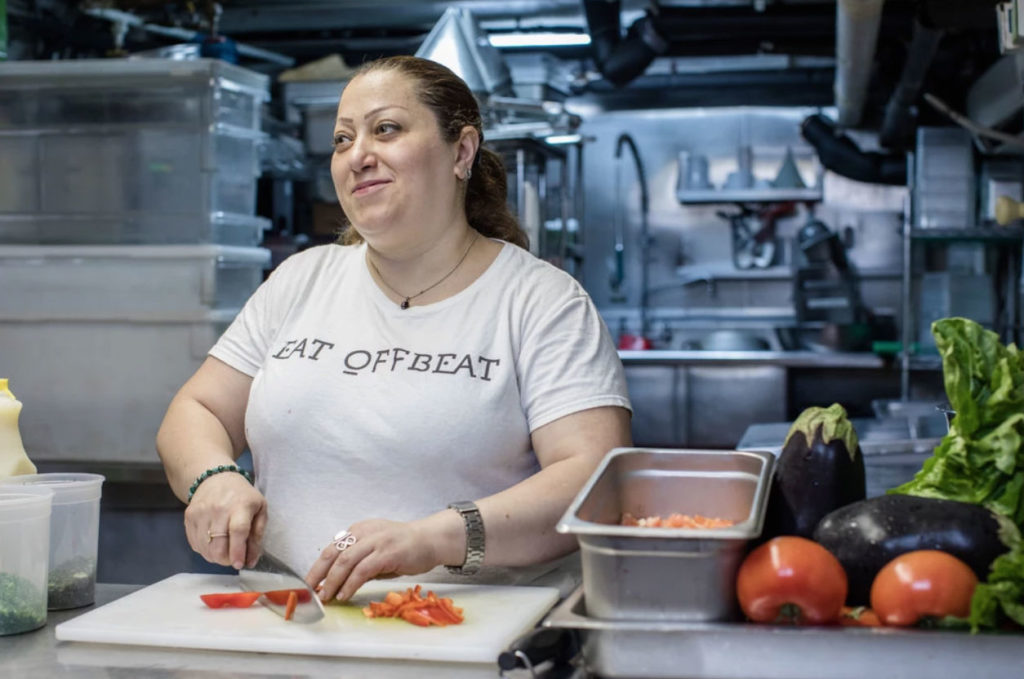 Woman cutting peppers in kitchen, wearing Eat Offbeat shirt.